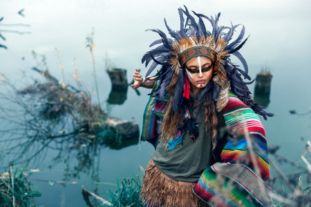 Beautiful girl dressed in native American clothes, with paint face camouflage and headdress with...