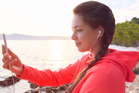 Active young woman listening to music and making selfie. Runner resting from routine exercise. Happy jogger relaxing at the beach after run and standing on the stones.の写真素材