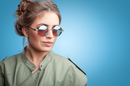 Close-up face portrait of young beautiful woman with perfect skin in round sunglasses looking up on blue background. Beauty face make-up.の写真素材