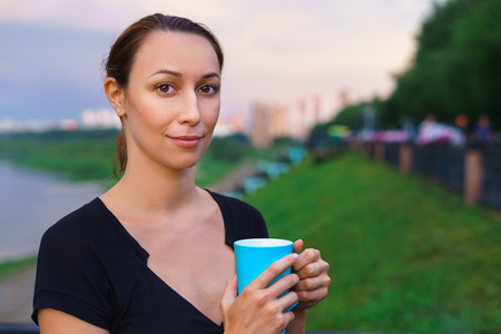 A girl stands on the embankment with cup of tea in his hands. The blue circle, the evening after the rain, in the street. Morning coffee outsideの写真素材