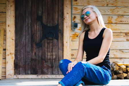 Girl sitting on a porch, in a wooden house. Young blonde with sunglasses. Smotrt aside, her hands folded in her kneesの写真素材