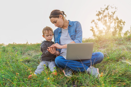 Mother and son sitting on the grass. Woman and baby are resting in nature, with a laptop on her knees. Boy interacts with her mother, the touch of hands. Wireless internet out of town.の写真素材
