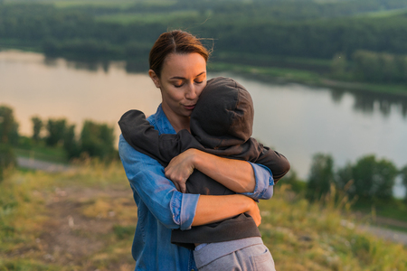 Mom hugging her son. Woman hugging a little boy outdoor. The concept of family in nature, unity with nature, gentle embrace. The landscape and the hill, river and forest. Sun going down. Augustの写真素材