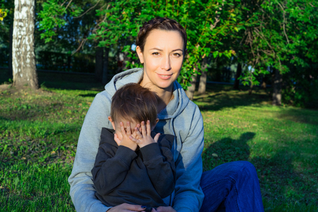 Mother and son sitting on the grass in the park. Family enjoying the outdoors. The kid in the arms of her mother. A boy covers his face with his hands. Shy. Sunny summer evening. A green lawn and trees.の写真素材