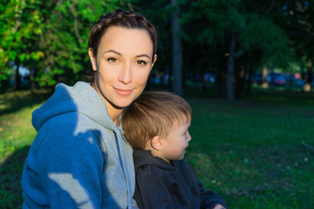 Mother and son sitting on the grass in the park. The kid in the arms of her mother. Sunny summer evening. A green lawn and trees.の写真素材
