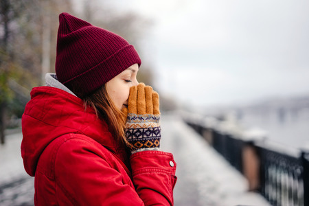 Girl presses the hands of her nose, his hands in mittens, cold autumn day, the first snow. Girl walks in the park in a red coat.の写真素材