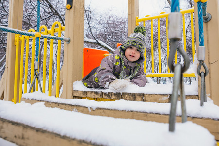 Boy is crawling on the playground in the park, winter day, snow on the playground, winter clothes in the childの写真素材