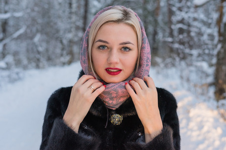Portrait of a young girl in a winter forest. Snow-covered trees, winter clothes, a coat and a scarf on her head. Make-up, red lips, blue eyesの写真素材