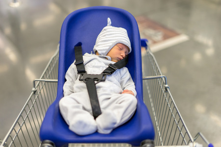 The little boy is in the chair on the trolley in the supermarket, the baby is sleeping in the store. A trip to the supermarket with a trolley for children.の写真素材