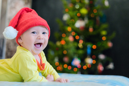 Little boy in a New Year's red hat is happy and smiles. Home comfort and warmth, and Christmas lights on a Christmas tree. Baby laughing and crawling on the bedの写真素材