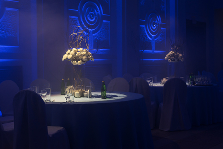 Table setting, wine glasses, piles. Bouquets of flowers in vases. Banquet room in a blue haze. Wedding decoration of the hall. Twilight and blue lighting, lamps and candles. White tablecloth. Layingの写真素材