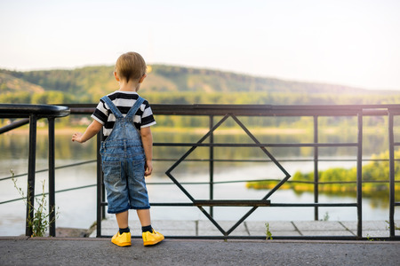 The boy stands on the edge of the ledge. The child walks along the alley off the coast. The railing closes the passage to the stairs. Cliff above the river beyond the fence.の写真素材