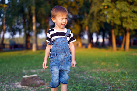 A child walks in the park, a denim overall. Evening sunny park. Green grass and trees. The boy jumps over the stumps.の写真素材