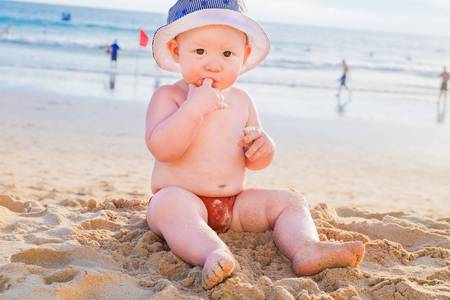 A kid with a finger in his mouth sits on the sand by the sea. A little boy in a panama on his head sits on the beach.の写真素材