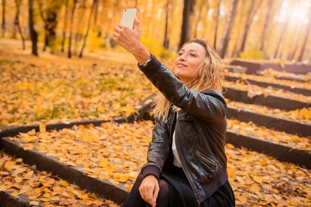 The girl is sitting on the stairs in the autumn forest. Brunette in a leather jacket with a phone in his hands, sms and internet surfing. Yellow leaf fall.の写真素材