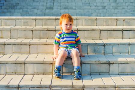 The boy is sitting on the steps. The red-haired baby sits on stone steps on a summer evening.の写真素材