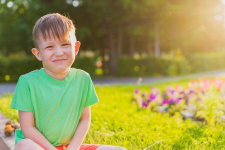 The boy sits grimaces and poses. Portrait of a preschooler. The boy sits in a park on a bed in the rays of the evening sun.の写真素材