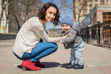 Mom teaches baby to walk on the street. Mom supports her son with her hands, teaches him to stand. A little boy learns to walk.の写真素材
