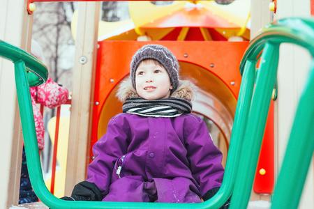 The boy plays on the playground in the winter. Cheerful baby is playing on the playground. Scarf,...