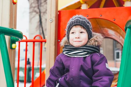 The boy plays on the playground in the winter. Cheerful baby is playing on the playground. Scarf, hat, gloves,の写真素材