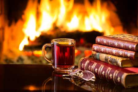 Glass mug of hot drink or alcoholic drink or mulled red wine and antique books in front of warm fireplace. Magical relaxed cozy atmosphere near fire. Autumn or winter conceptの写真素材