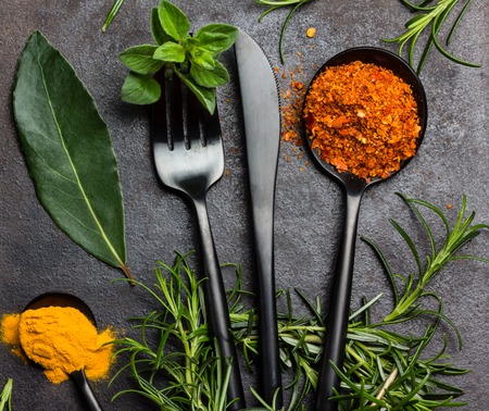 Food background with fresh herbs - rosemary and oregano, dryed spices - chili pepper, turmeric curcuma and black steel cutlery - knife spoon fork, tea spoon on black background. Top view. Copy spaceの写真素材