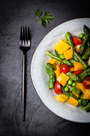 Mediterranean vegetarian salad. Vegan salad with asparagus, cherry tomatoes, yellow bell pepper on gray plate on black slate stone background. Vegetarian or healthy food. Top viewの写真素材