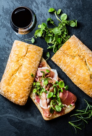 Ciabatta sandwich with jamon ham serrano paleta iberica, arugula, rosemary and glass of red wine on stone slate black background. Top viewの写真素材