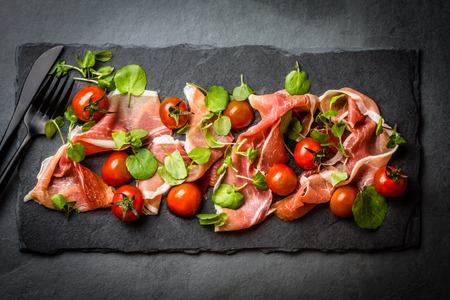 Salad with ham jamon serrano, cherry tomatoes, arugula on black stone slate board, grey background. Top viewの写真素材