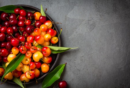 Ripe red and yellow cherry with leaves on black plate plate, grey stone background. Top viewの写真素材