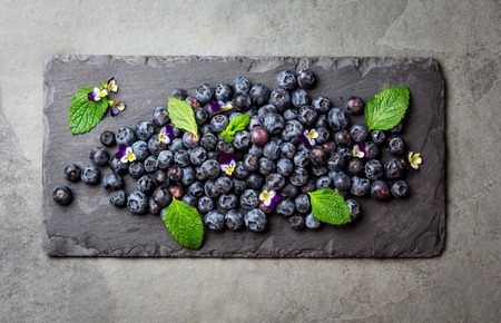 Berry fresh blueberries with fresh mint leaves and edible flowers on black slate board. Stone gray background, healthy food, diet, detox concept. Top viewの写真素材