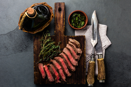 Slices medium rare beef steak with herb sauce, bottle of wine, vintage cutlery, slate backgroundの写真素材