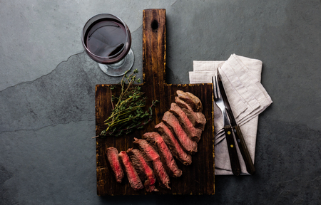 Slices of beef medium rare steak on wooden board, glass of red wine on slate backgroundの写真素材