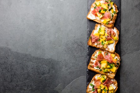 Toasts with cream cheese, ham jamon serrano and mango served on black slate board with red wine, top view, gray slate background, copy spaceの写真素材