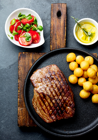 Grilled beef steak served on cast iron plate with tomato salad and potatoes balls. Barbecue, bbq meat beef tenderloin. Top view, slate background.の写真素材