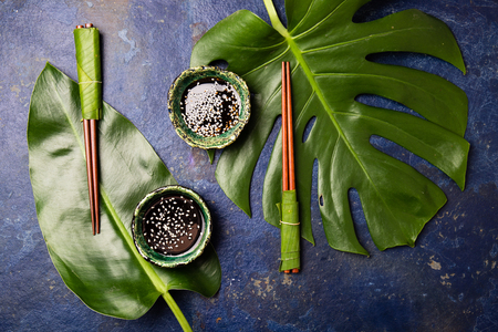 Asian food concept. Chopstick and soysauce soy sauce with white sesame on blue background with tropical leaves . Top viewの写真素材