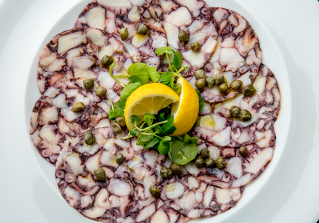 OCTOPUS CARPACCIO. Seafood Raw octopus slices with olive oil, lemon and capers on white plate. Top view. Gray stone background.の写真素材