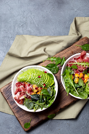 Low carbs bowl. Fresh salad with green spinach, rucola, avocado an ham serrano in white bowl, gray background, top view.の写真素材