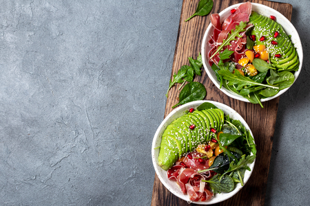Low carbs bowl. Fresh salad with green spinach, rucola, avocado an ham serrano in white bowl, gray background, top view.の写真素材