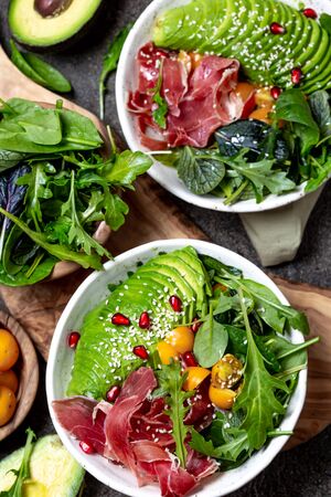 Low carbs bowl. Fresh salad with green spinach, rucola, avocado an ham serrano in white bowl, gray background, top view.の写真素材