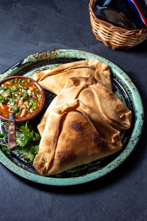 Tipical baked empanadas de pino with pebre sauce on vintage try, black stone background. Traditional chilean food for independence day partyの写真素材