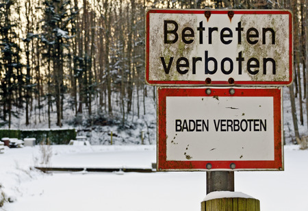 signs labelled \"bathing forbidden\" and \"Keep off!\" at a forest pond in winterの写真素材