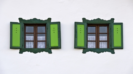 two wooden windows with green fringes and shutters at a white wall