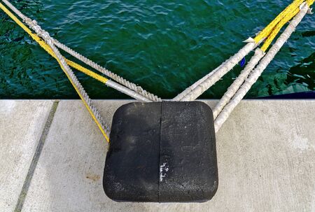 black quadratic bitt and white and yellow mooring ropes at a quay wall with view on the green waterの写真素材