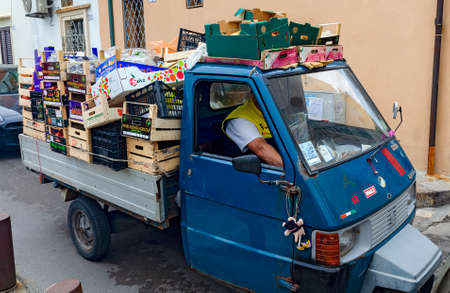 market driver with three-wheeled motor car high loaded with boxes and crates in a narrow street of...