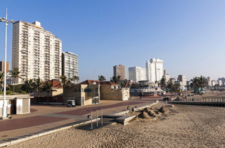 DURBAN, SOUTH AFRICA - JUNE 24, 2016: Early morning empty beach front paved promenade and two unknown  sand castle builders against Golden Mile city skyline in Durban, South Africaのeditorial素材