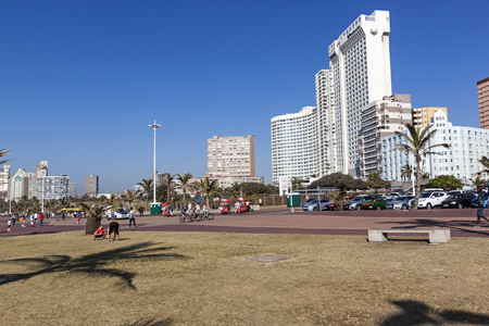 DURBAN, SOUTH AFRICA - JUNE 26, 2016:Grass verge and  many unknown people on  early morning walking and cycling  on paved promenade on Golden Mile beach front against city skyline in Durban, South Africaのeditorial素材