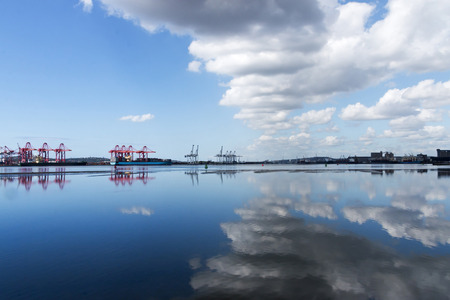 Harbor at low tide with cloudy blue sky reflecting in water and container vessels cranes and city skyline in backgroundの写真素材