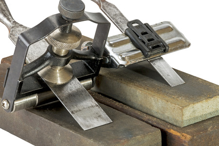 Studio shot of two chisels clamped in angle guide jigs resting on grinding whetstones on white...