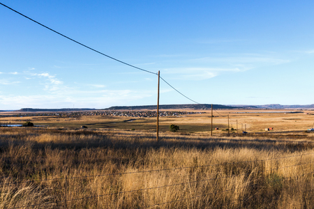 Fenced autumn colored dry grass against rural country residential settlement and blue sky horizon in South Africaの写真素材
